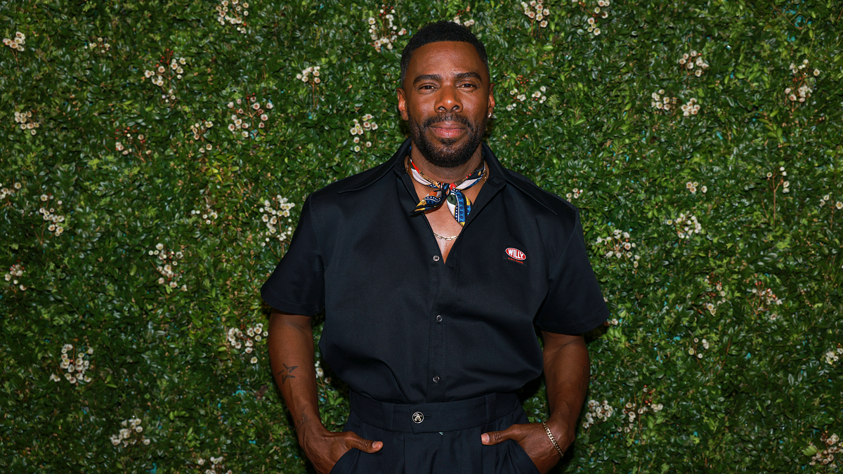 Colman Domingo wears a stylish black outfit with a neck scarf, posing in front of a green floral backdrop