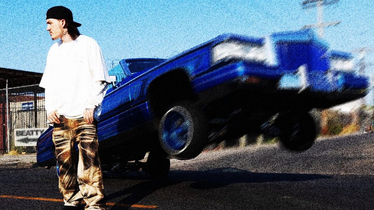 Person in streetwear and a backwards cap stands near a blue lowrider car lifting its front wheels in a street setting. Image is categorized as Music