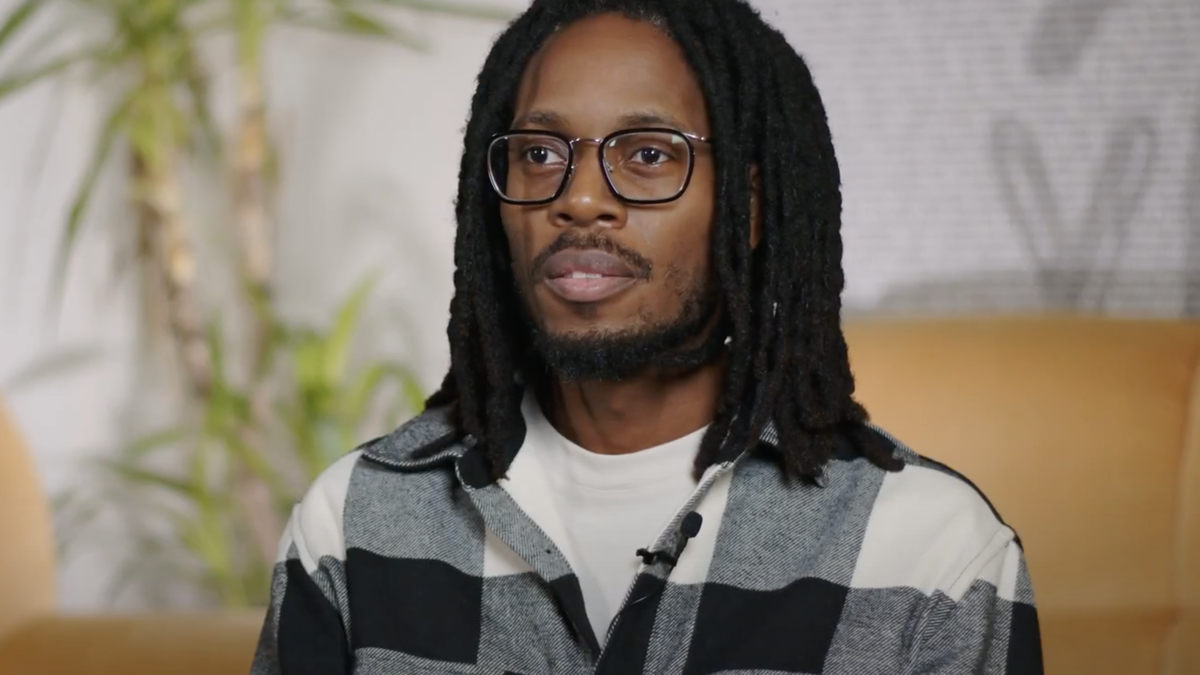 A man with glasses, wearing a black-and-white checkered shirt, is seated indoors in a setting with plants and a light background