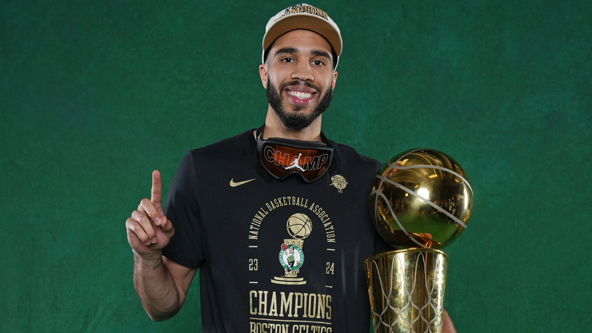 Jayson Tatum holding an NBA Championship trophy and wearing a "Champions" t-shirt, cap, and championship goggles, smiling and pointing upward with one finger