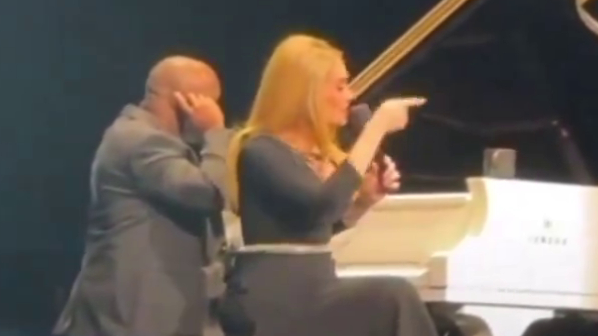 Adele performs on stage, singing into a microphone while seated at a white piano. Backup musician stands nearby