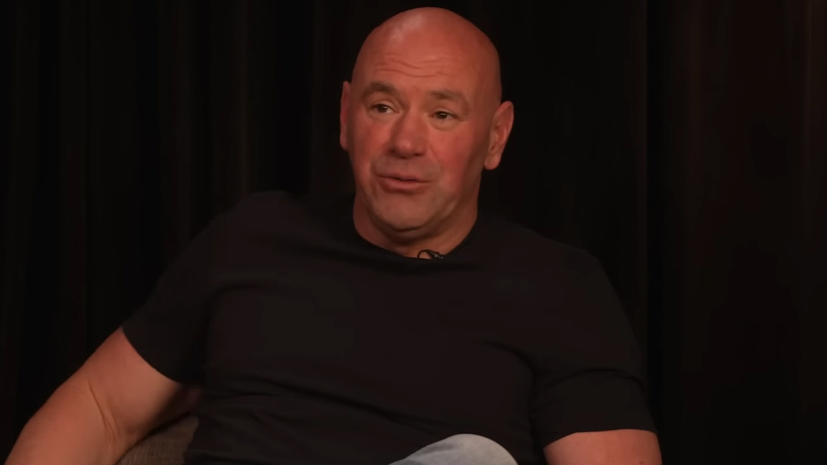 Dana White sits wearing a casual black t-shirt and jeans, discussing in an interview setting with a dark background
