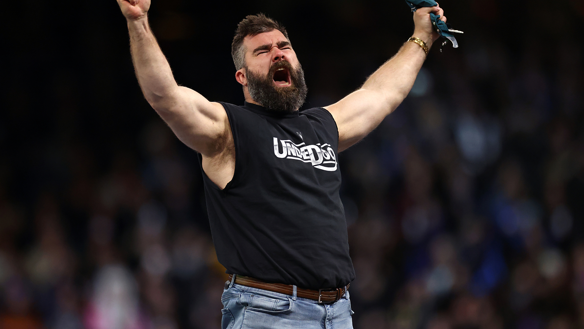Jason Kelce celebrates passionately with both arms lifted, wearing a sleeveless black shirt and jeans, in a sports arena