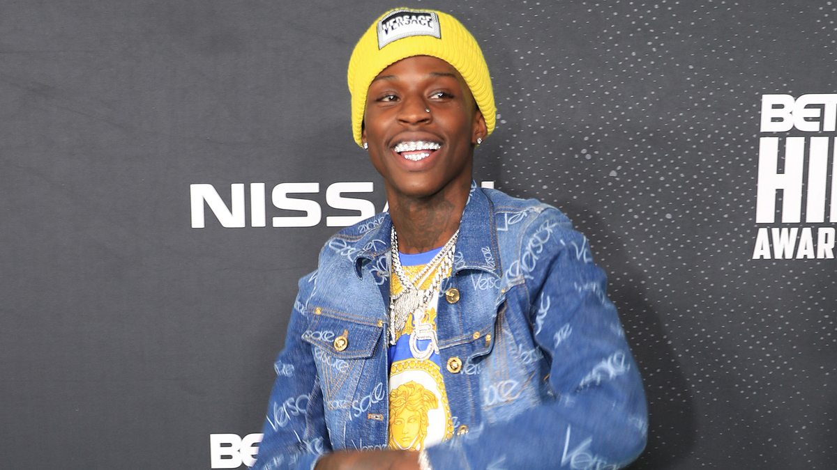 A person is smiling while wearing a yellow beanie and a blue denim jacket with text patterns, standing at the BET Hip Hop Awards 2019