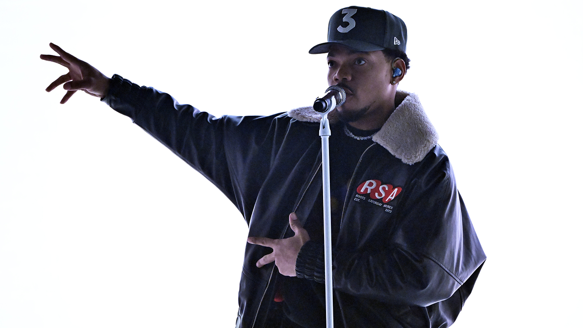 Chance the Rapper performing on stage, wearing a dark jacket with fur trim and a baseball cap with the number "3" on it