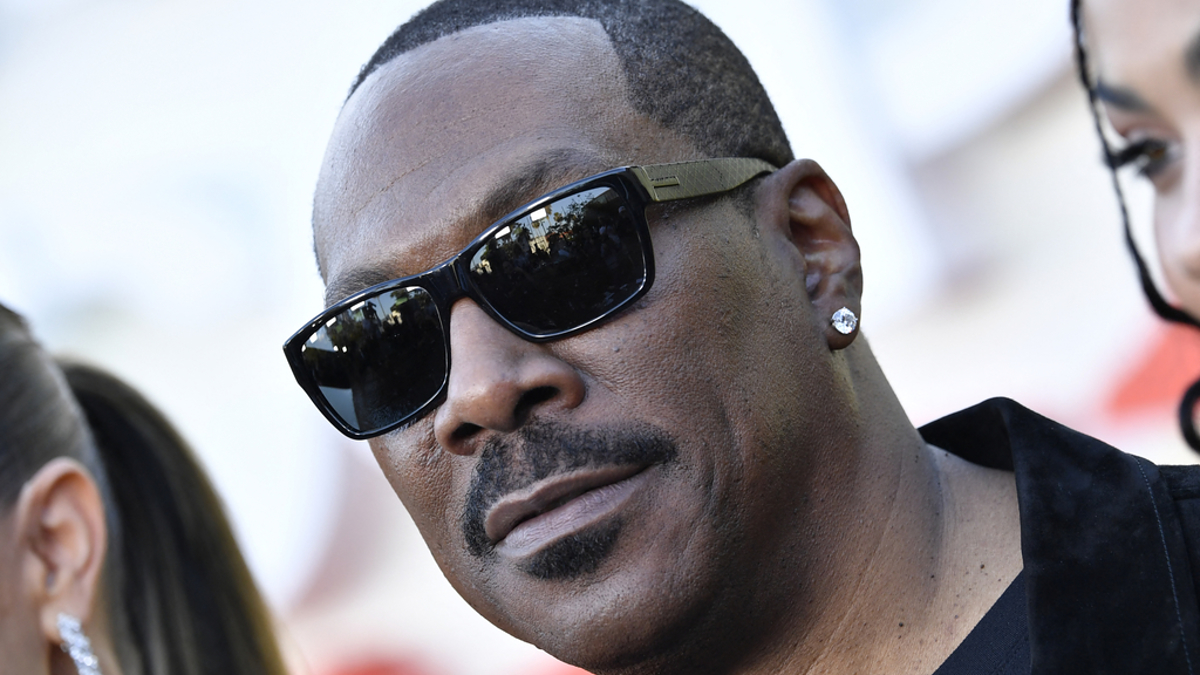 Eddie Murphy wearing sunglasses and a black jacket, posing on a red carpet