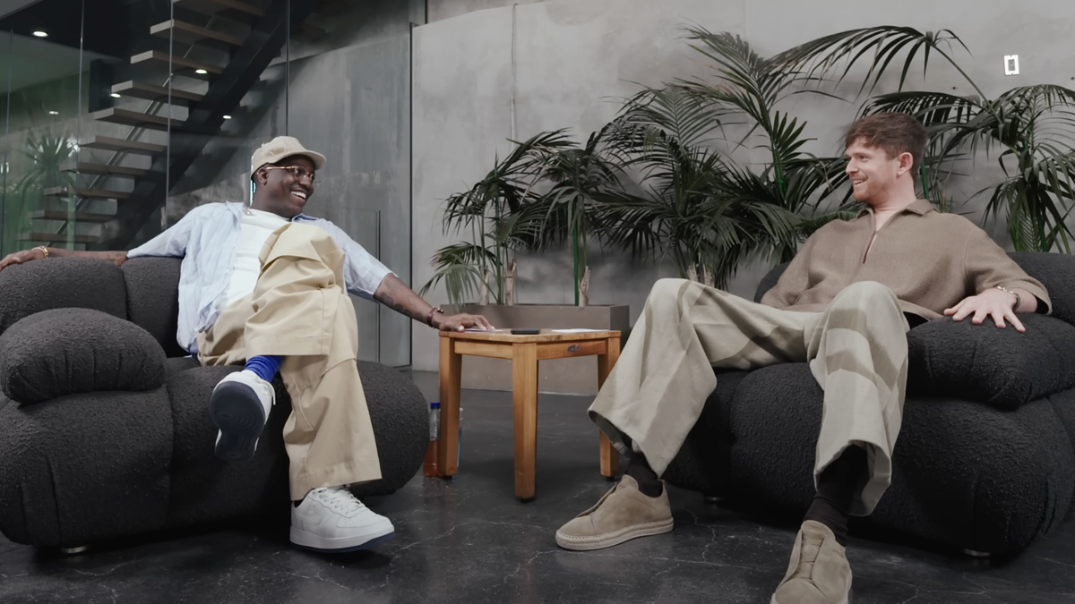 Lil Yachty and James Blake sit on black armchairs facing each other, smiling and talking in a modern setting with a wooden table and plants in the background