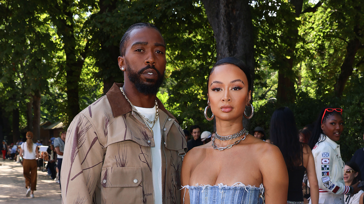 Orlando Scandrick and Draya Michele pose outdoors, with Orlando wearing a jacket and Draya in a strapless dress, surrounded by trees and people in the background