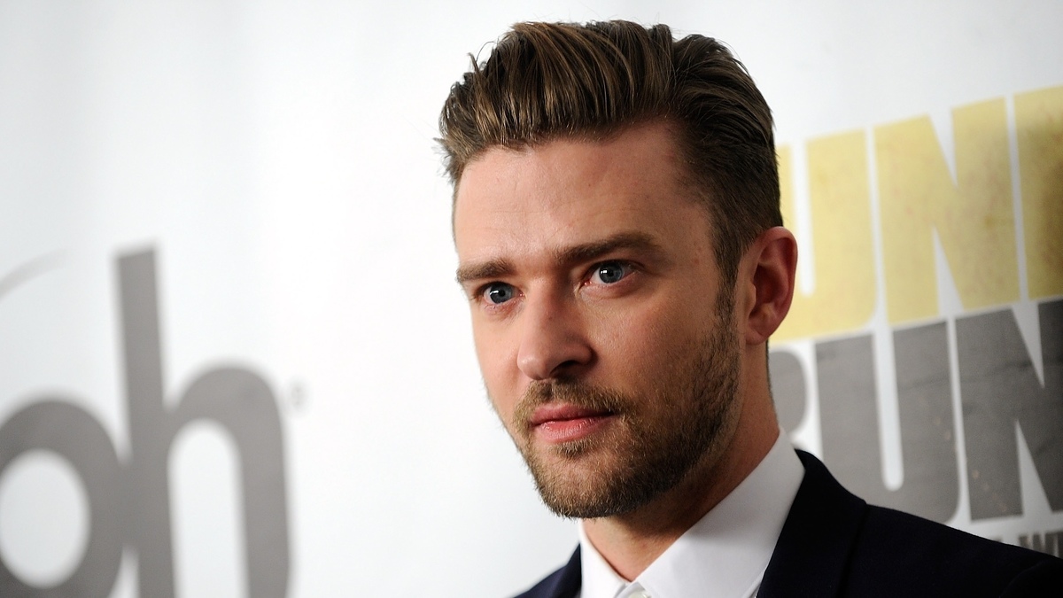 Justin Timberlake in a suit at a media event with a backdrop displaying partially visible text