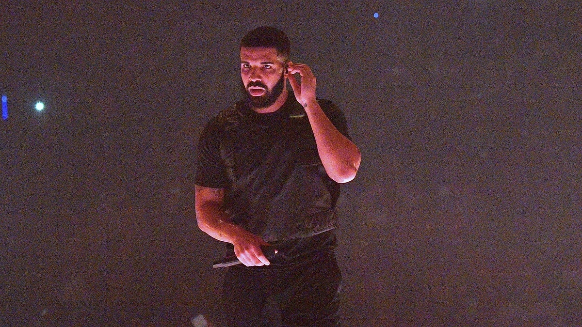 Drake, wearing a casual black outfit, performing on stage while holding a microphone to his side. He appears focused and is adjusting his earpiece