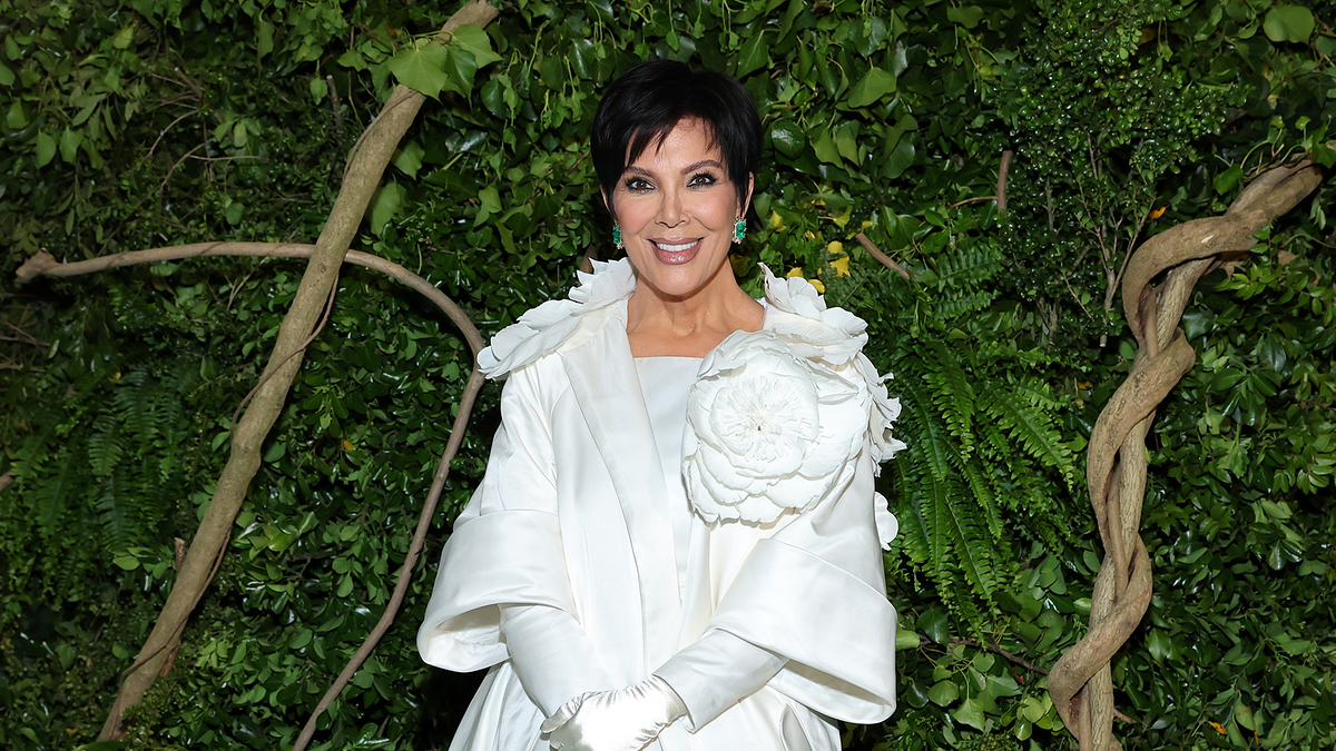 Kris Jenner stands in front of lush greenery, wearing an elegant, white coat with a large flower embellishment on the shoulder