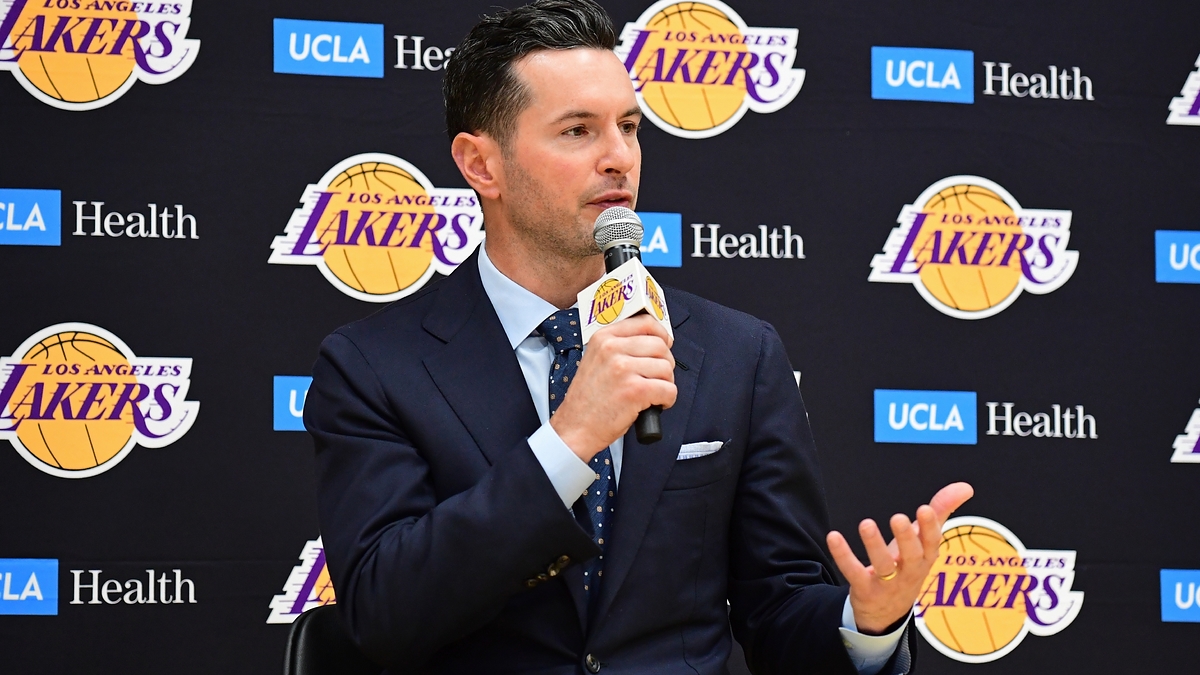 J.J. Redick in a press conference, speaking into a microphone while in a dark suit and tie, seated in front of a Los Angeles Lakers backdrop