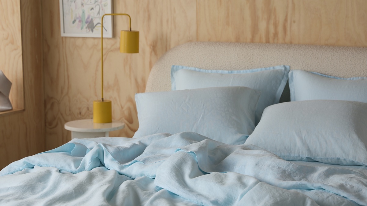 A neatly made bed with light blue sheets and pillows, a small white side table with two yellow lamps, and artwork hanging on a wooden wall in the background