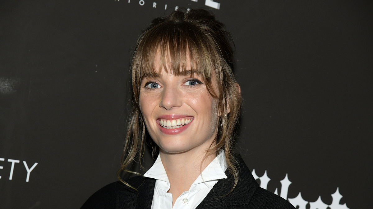 A person with bangs and a smile, dressed in formal attire with a white shirt and a dark jacket, standing against a backdrop with indistinct text