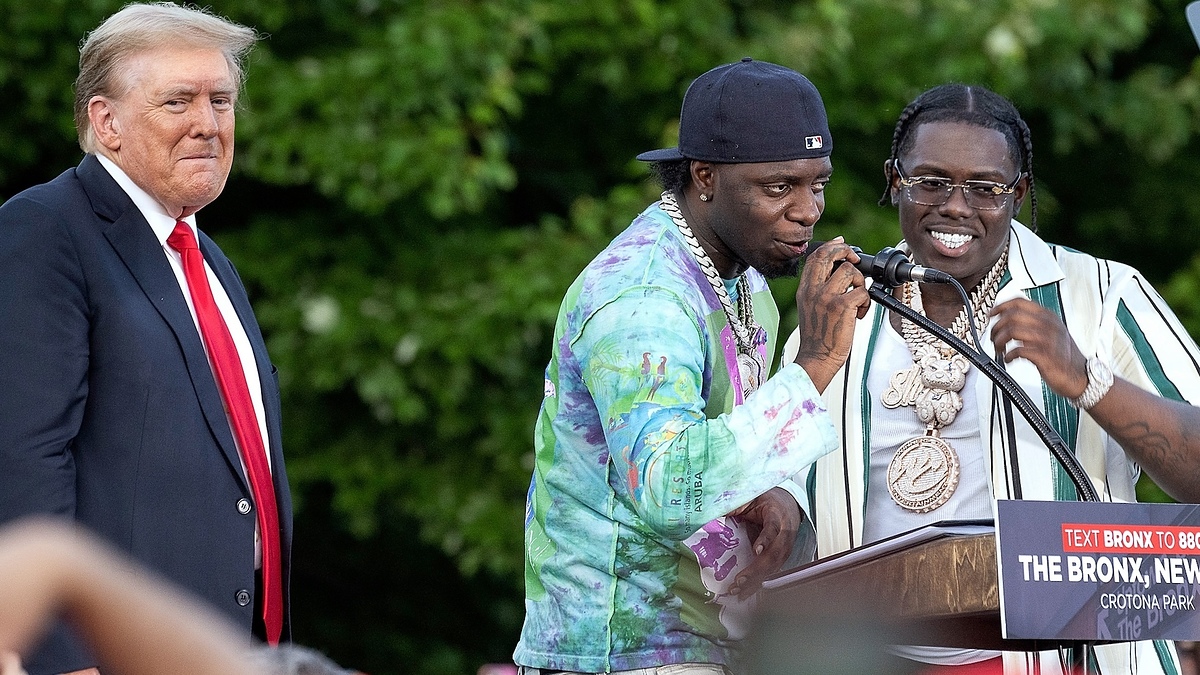 Donald Trump stands next to rappers Fresha and Symphony at a podium during an outdoor event. Fresha and Symphony are speaking into microphones