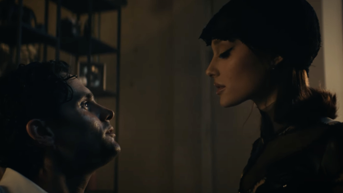Two individuals in a dimly lit room, one sitting and looking up, the other standing and looking down. Set in a dramatic scenario. Names unknown