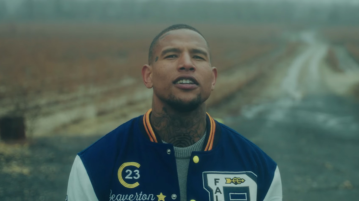 Darren Waller stands outdoors, wearing a letterman jacket with patches, including the letter 'B' and the number '23'. The background shows a path through a field