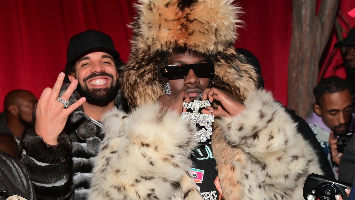 Drake smiles and throws a hand sign while standing behind Lil Yachty, who wears a large fur hat and coat, and holds a diamond-encrusted medallion