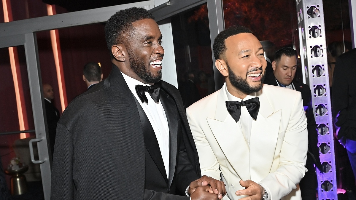 Sean Combs and John Legend are smiling and shaking hands at a formal event. Sean is wearing a black suit with a cape, and John is in a white tuxedo