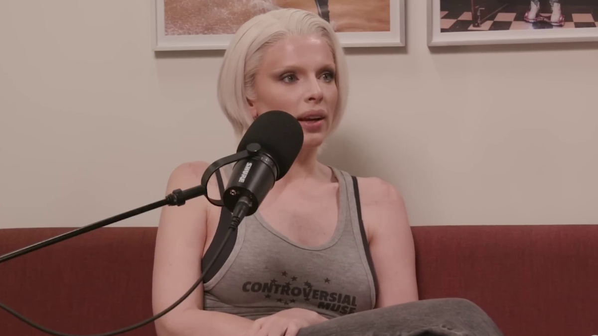 Julia Fox with short blonde hair, tank top that says "CONTROVERSIAL MUSE," speaking into a microphone while sitting on a couch