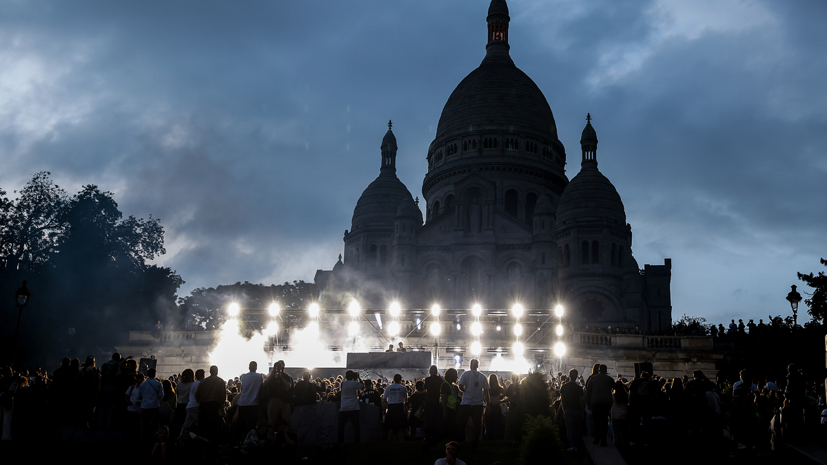 A large crowd gathers in front of a well-lit Sacré-Cœur Basilica during twilight, creating a dramatic and lively atmosphere