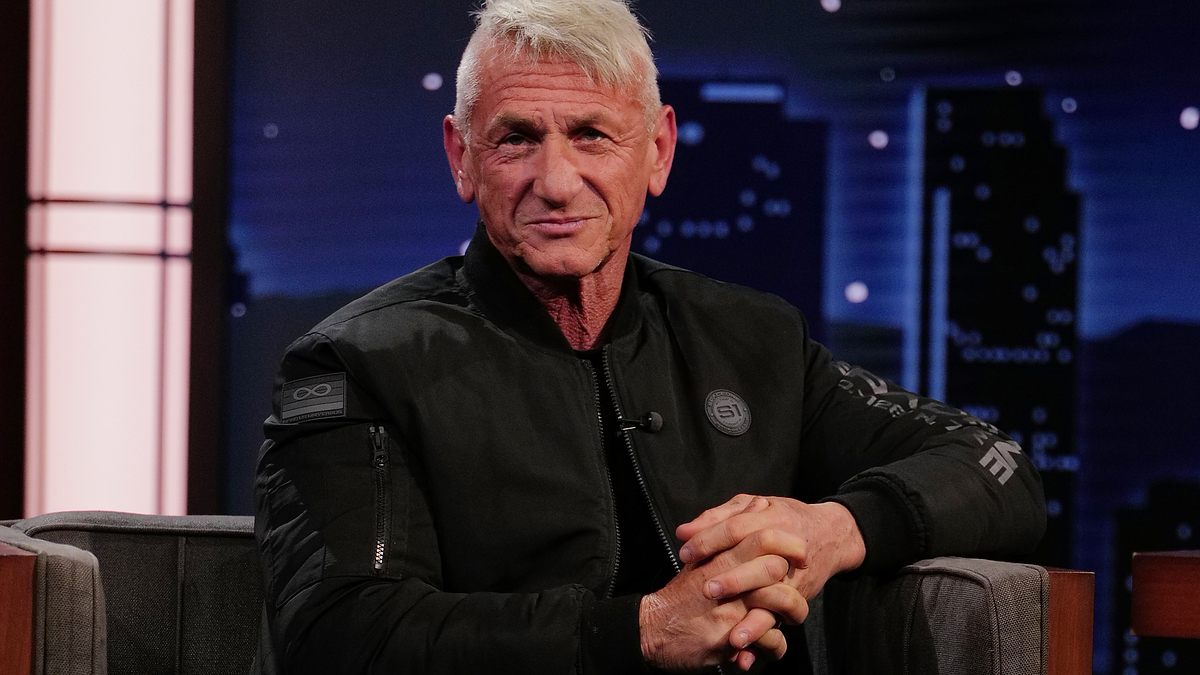 Sean Penn sits on a talk show set, wearing a black jacket and grey pants with white sneakers