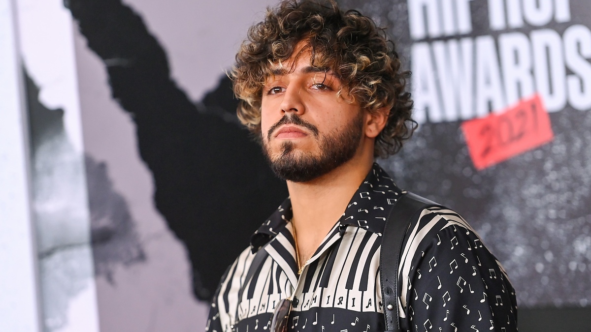 A man with curly hair and a beard stands at the 2021 Hip Hop Awards, wearing a patterned shirt with musical notes and piano keys. Names are not known