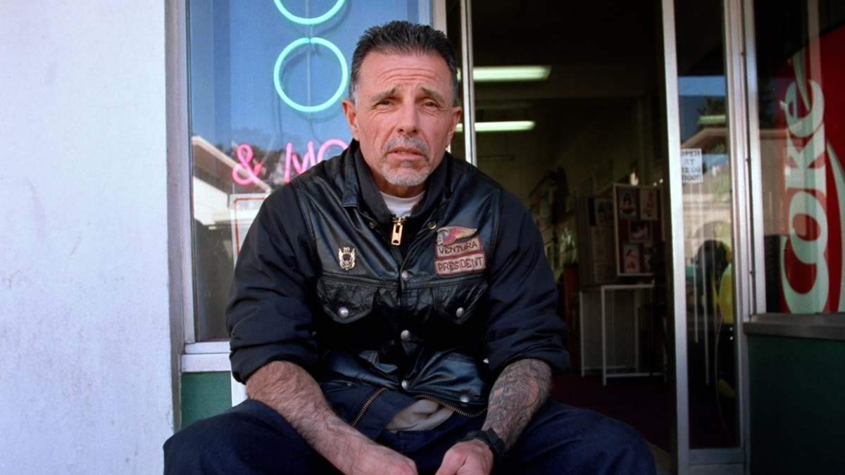 A man sits outside a shop wearing a leather jacket with patches, including one that reads "President." Neon signs are visible in the background