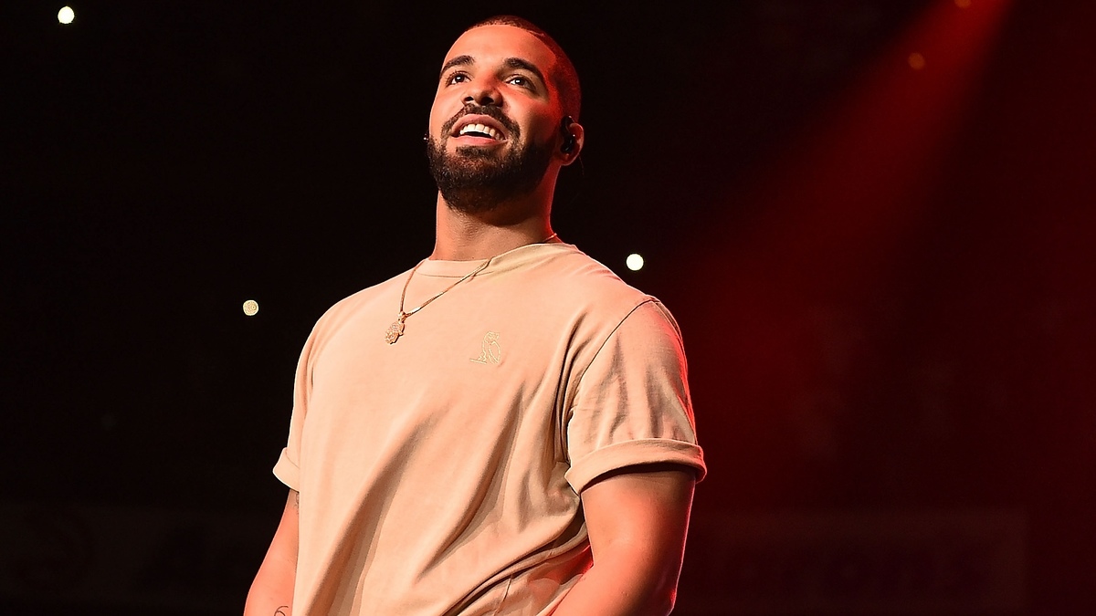 Drake smiles on stage, wearing a casual short-sleeved shirt and necklace