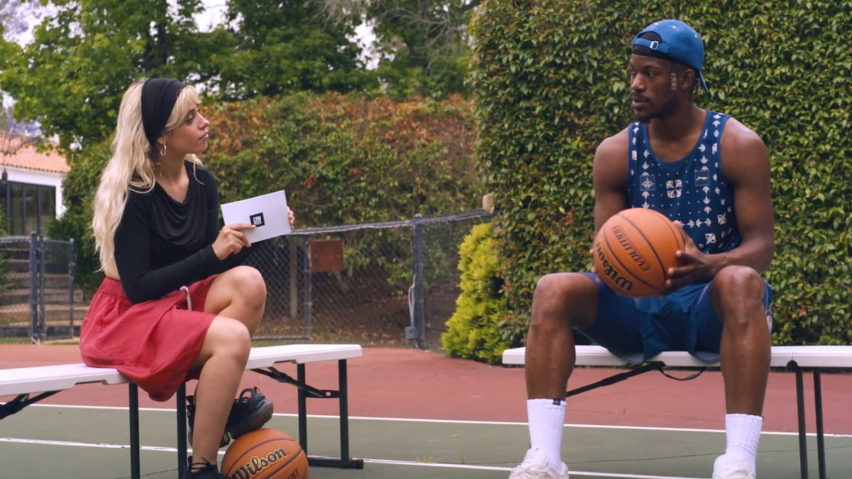 Camila Cabello holding a card with a microphone interviews Jimmy Butler in a sleeveless jersey and cap. Both hold basketballs while sitting on benches on an outdoor court