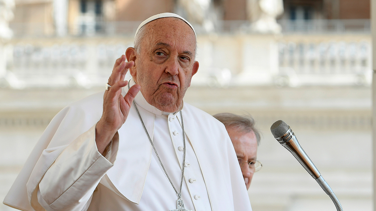 Pope Francis speaking into a microphone, wearing traditional papal attire with a cross necklace and white cassock, raising his right hand