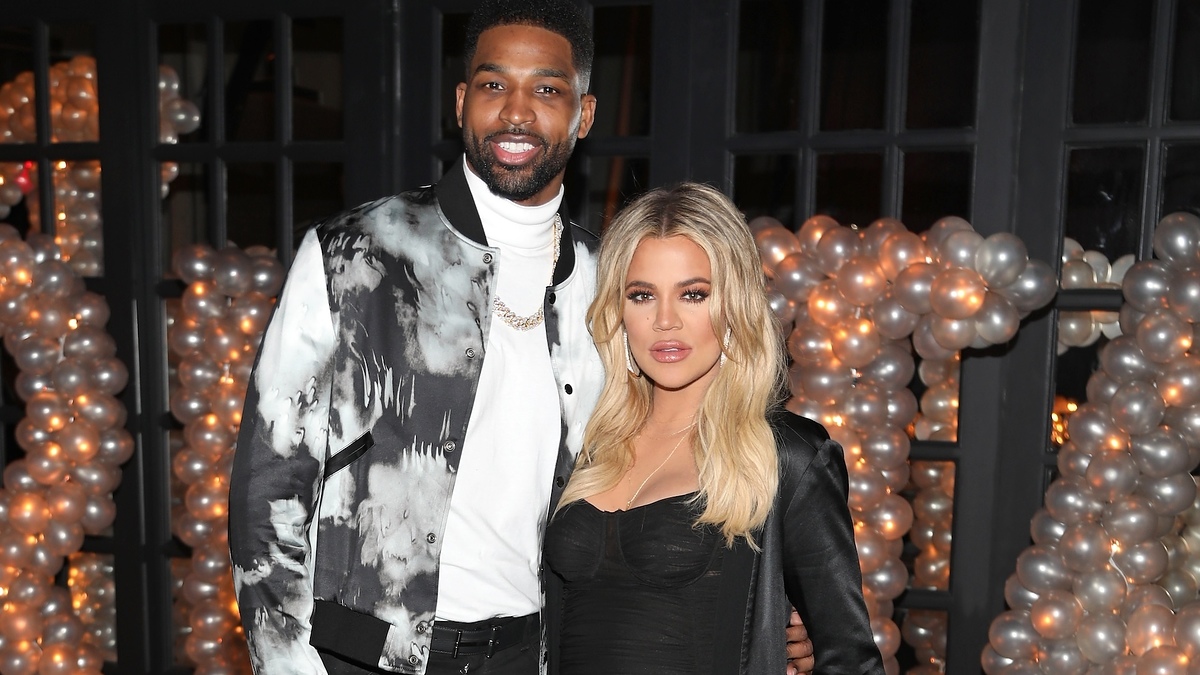 Khloé Kardashian in a fitted dress and Tristan Thompson in a patterned jacket pose together at an event with a balloon backdrop