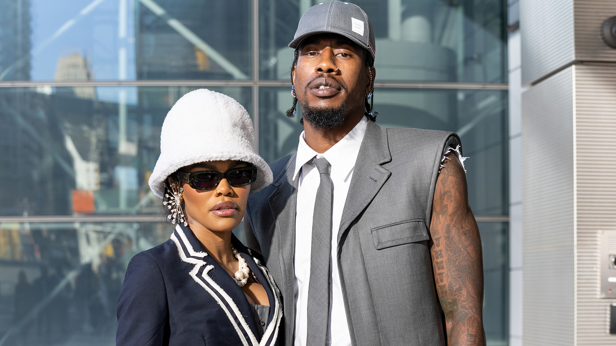 Teyana Taylor and Iman Shumpert pose together. Teyana wears a chic outfit with a white hat and large sunglasses. Iman is dressed in a suit and baseball cap