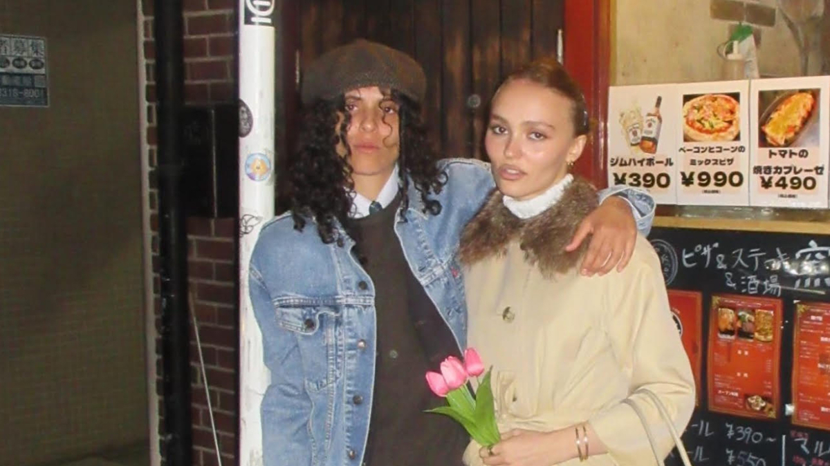 Lily-Rose Depp and 070 Shake pose together, with Ash in a denim jacket and beret, and Lily-Rose holding pink tulips while wearing a beige coat