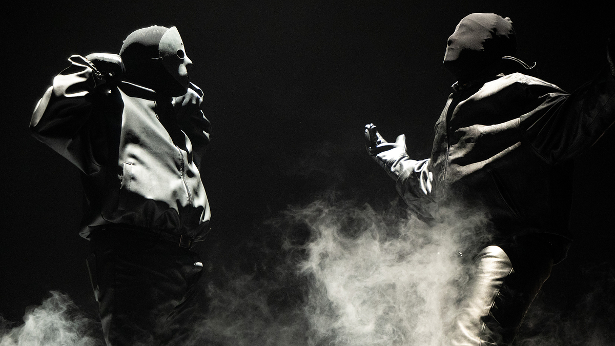 Two masked individuals in heavy jackets and pants perform on a foggy stage