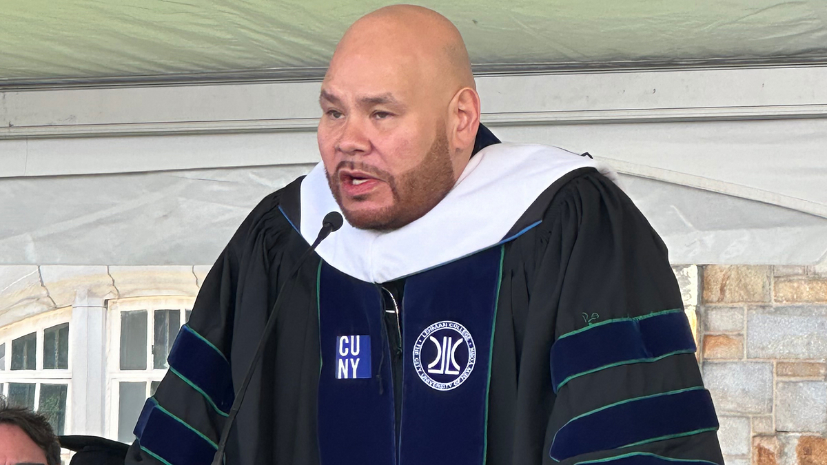 Fat Joe delivering a commencement speech at a graduation ceremony, wearing a traditional academic gown and hood on stage