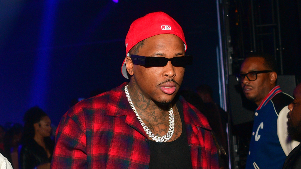 Hip hop artist YG, wearing a red cap and a red plaid shirt with a chain necklace, attends an event. Two other unidentified individuals are in the background