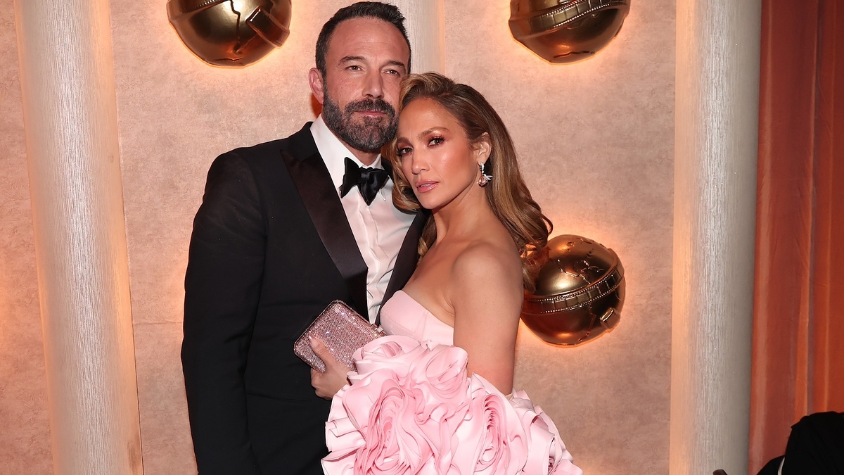 Ben Affleck and Jennifer Lopez pose together in formal attire, with Jennifer wearing a dramatic, floral-themed dress and holding a sparkling clutch