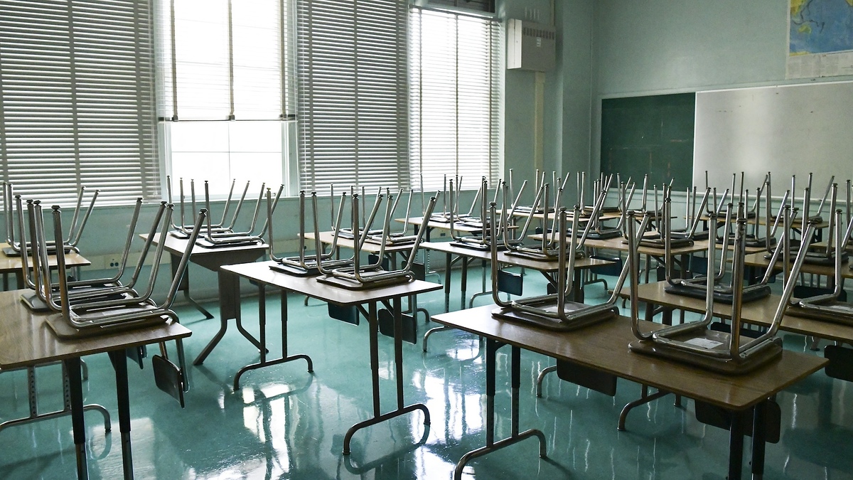 Desks with chairs stacked upside down in an empty classroom with sunlight coming through the window blinds
