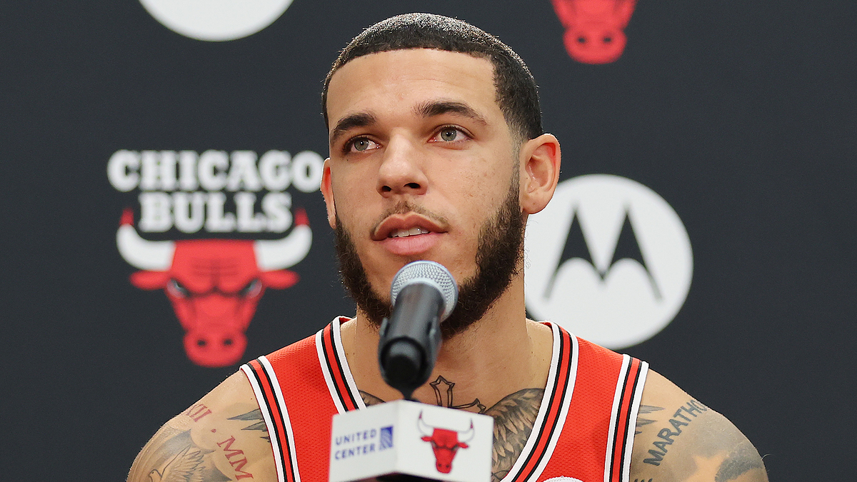 Lonzo Ball speaks at a Chicago Bulls press conference wearing team jersey, microphone in front of him. Bulls logo and sponsor logos are in the background
