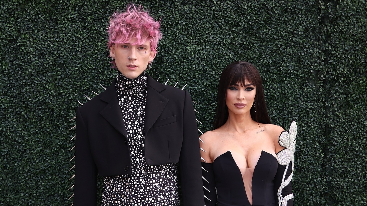 Machine Gun Kelly and Megan Fox stand together; Machine Gun Kelly wears a black outfit with metallic spikes and a polka dot shirt, while Megan Fox wears a fitted black dress