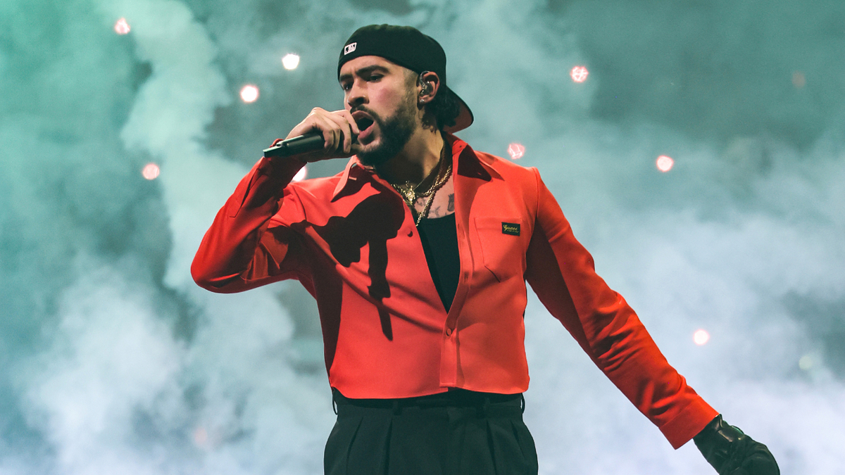 Bad Bunny performs on stage wearing a red shirt and a backward black cap, holding a microphone, surrounded by smoke effects