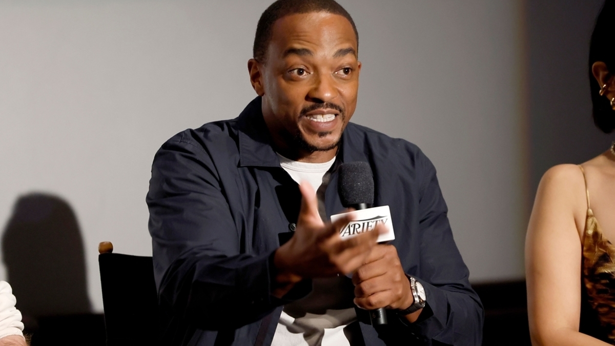 Anthony Mackie speaking into a microphone while gesturing with his hand. He is wearing a casual jacket and sitting in a chair