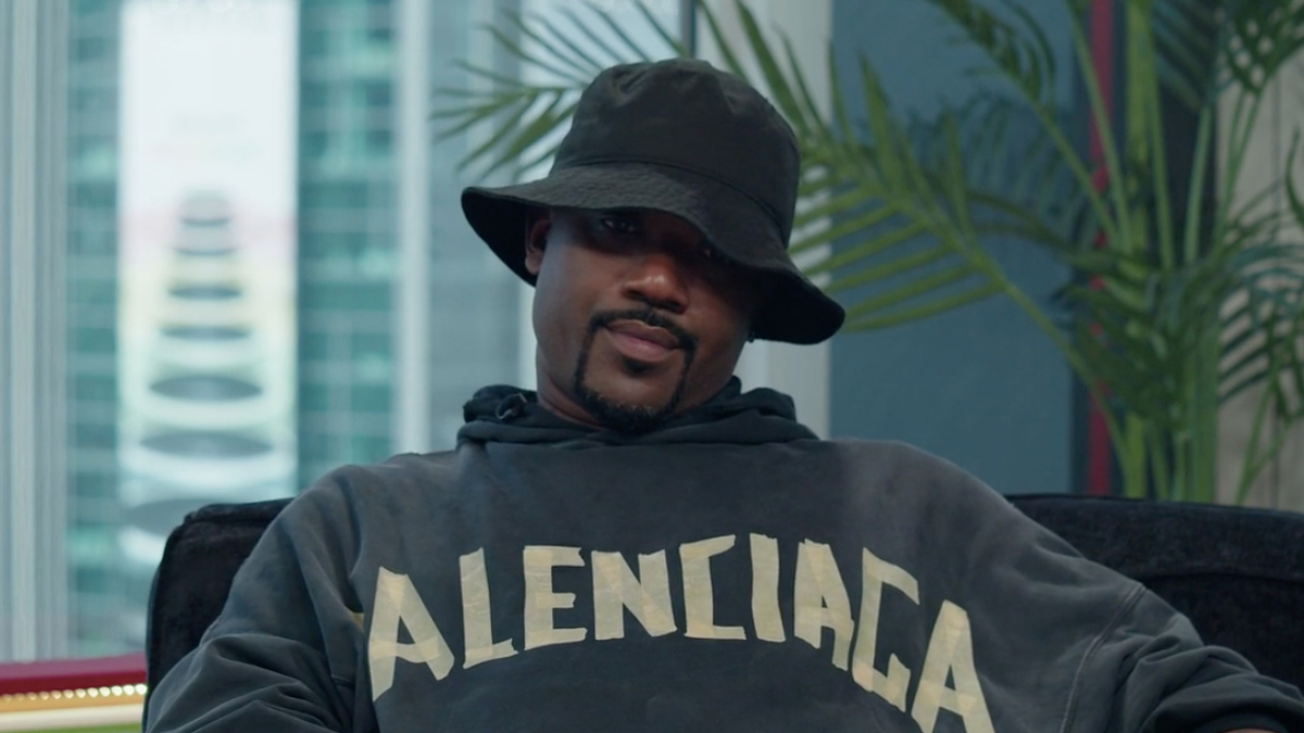 Ray J wearing a Balenciaga hoodie and a black bucket hat, seated indoors with a plant and buildings visible in the background
