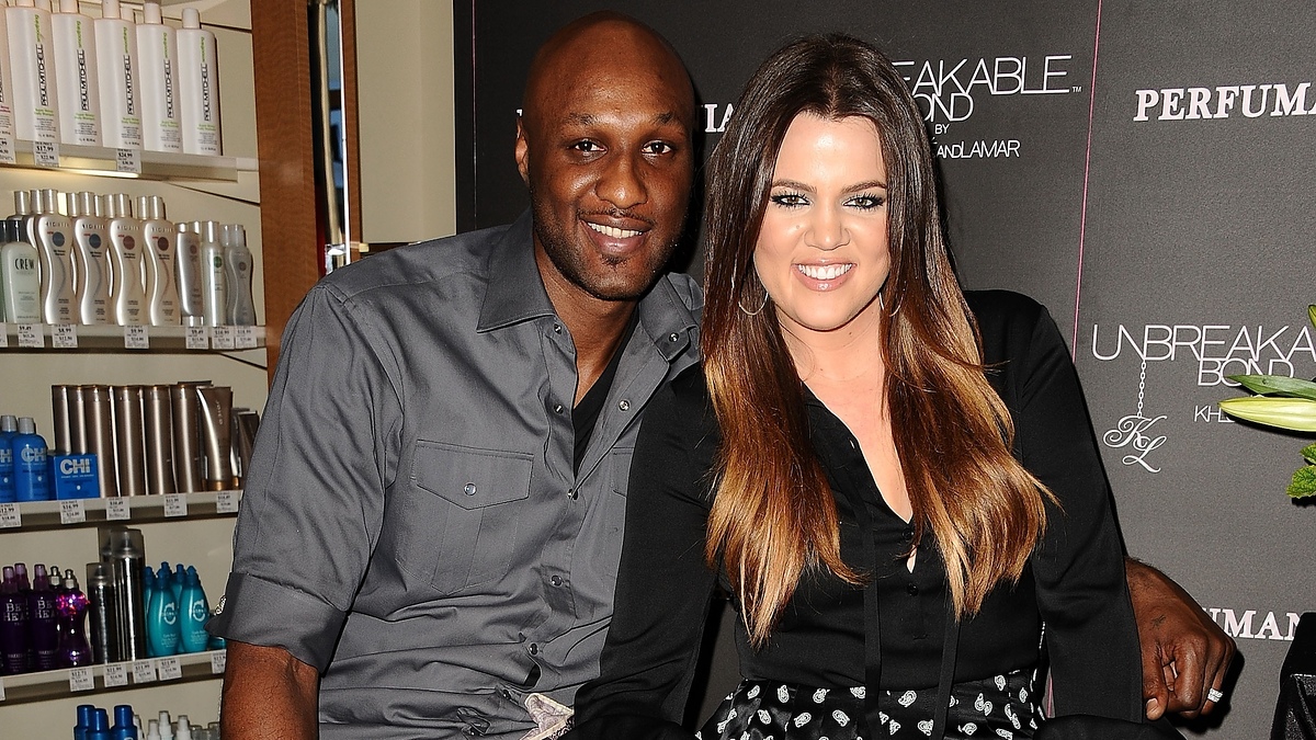 Lamar Odom and Khloé Kardashian pose together in front of perfume displays. Khloé wears a black blouse and patterned skirt