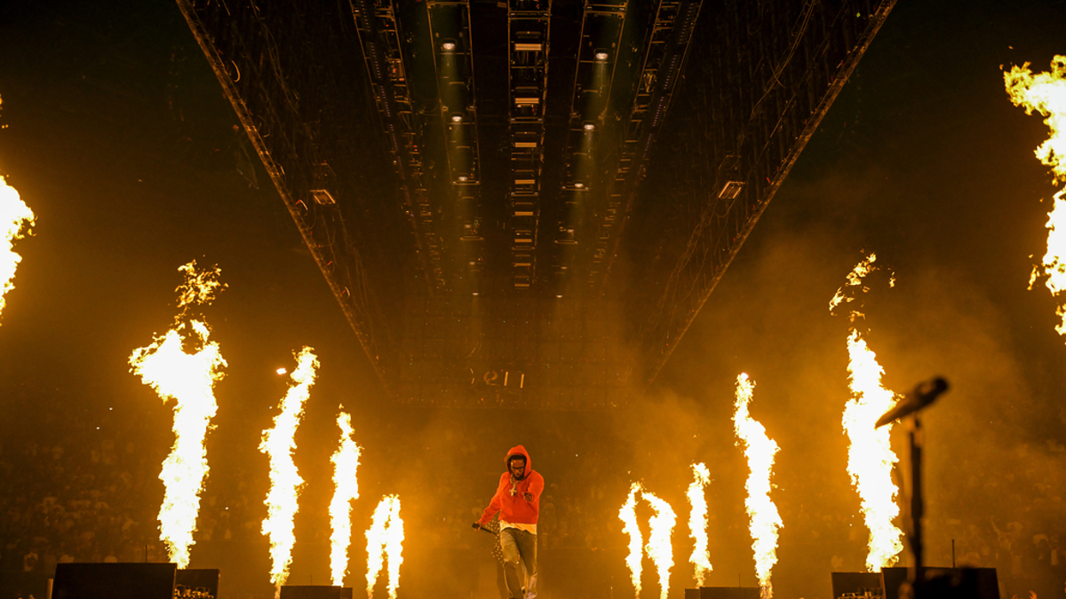 Musician performs on stage amid tall flames and a large crowd at a concert, wearing a hooded sweatshirt