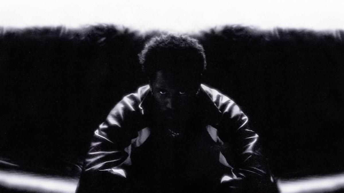 A person with a serious expression, seated with arms on knees, in a dimly lit setting