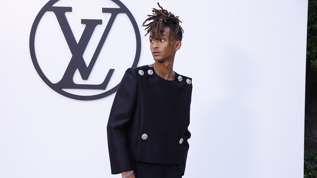 Jaden Smith on a red carpet, wearing a stylish black suit with embellishments, standing in front of a large Louis Vuitton logo