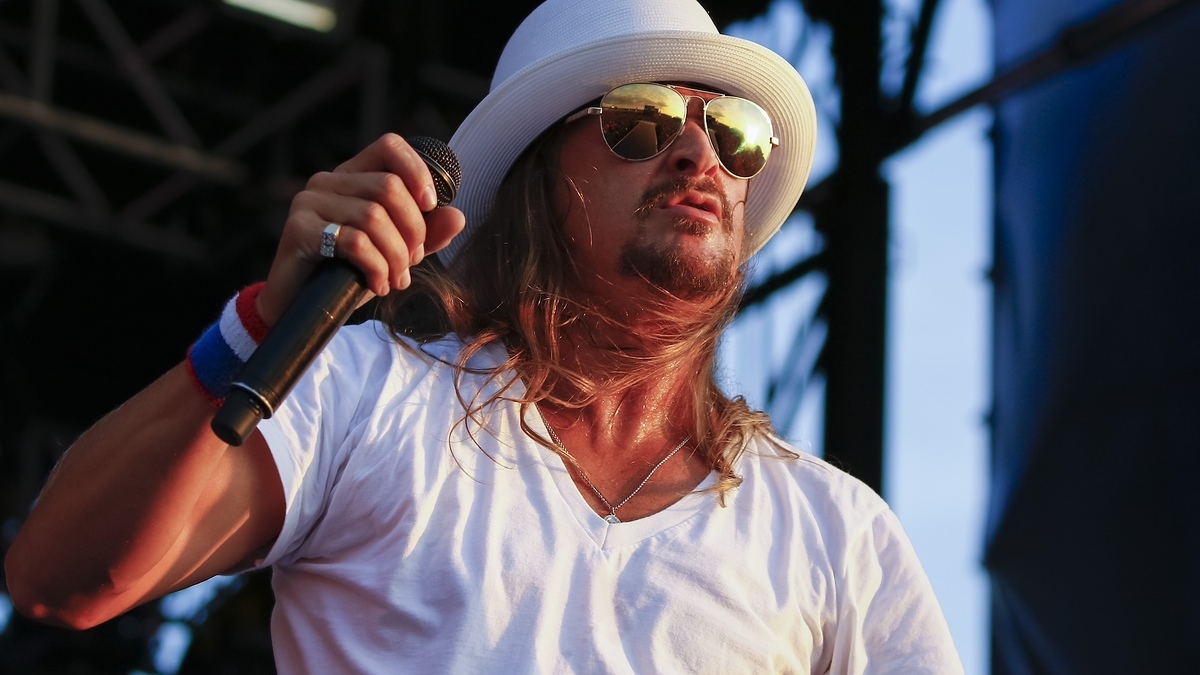 Kid Rock performing on stage, wearing a white t-shirt, white fedora, aviator sunglasses, and a silver necklace, holding a microphone