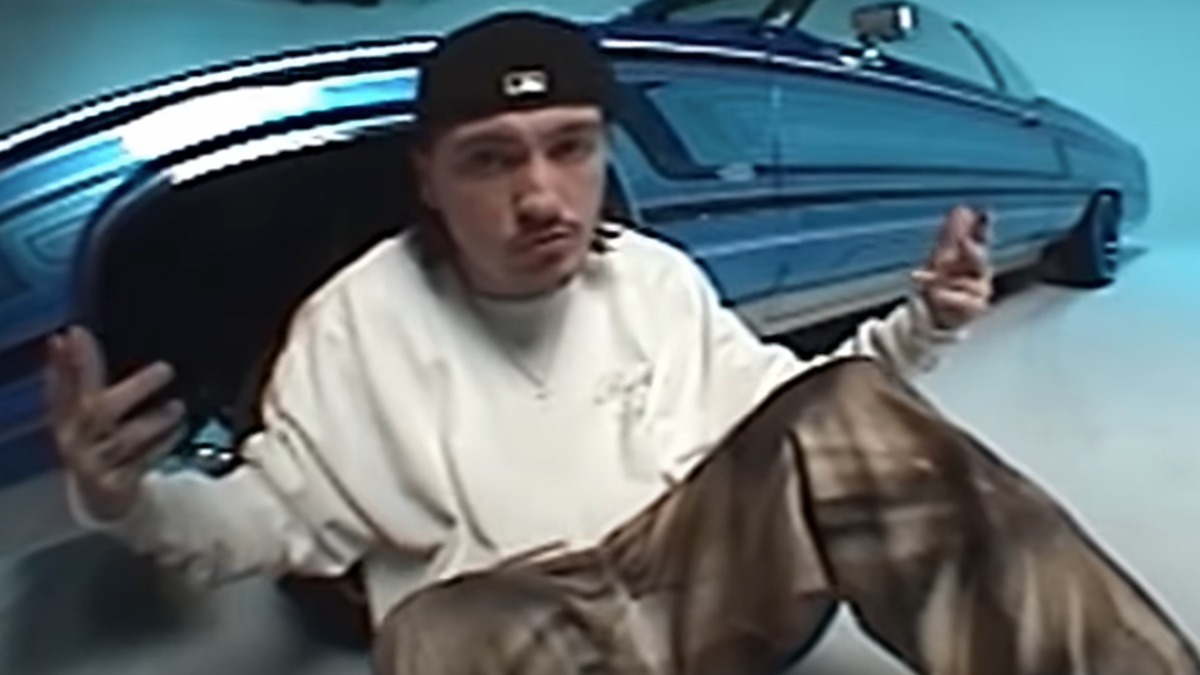 Man sitting in front of blue car, wearing a white long-sleeve shirt, baggy plaid pants, and a backwards cap in a music video setting